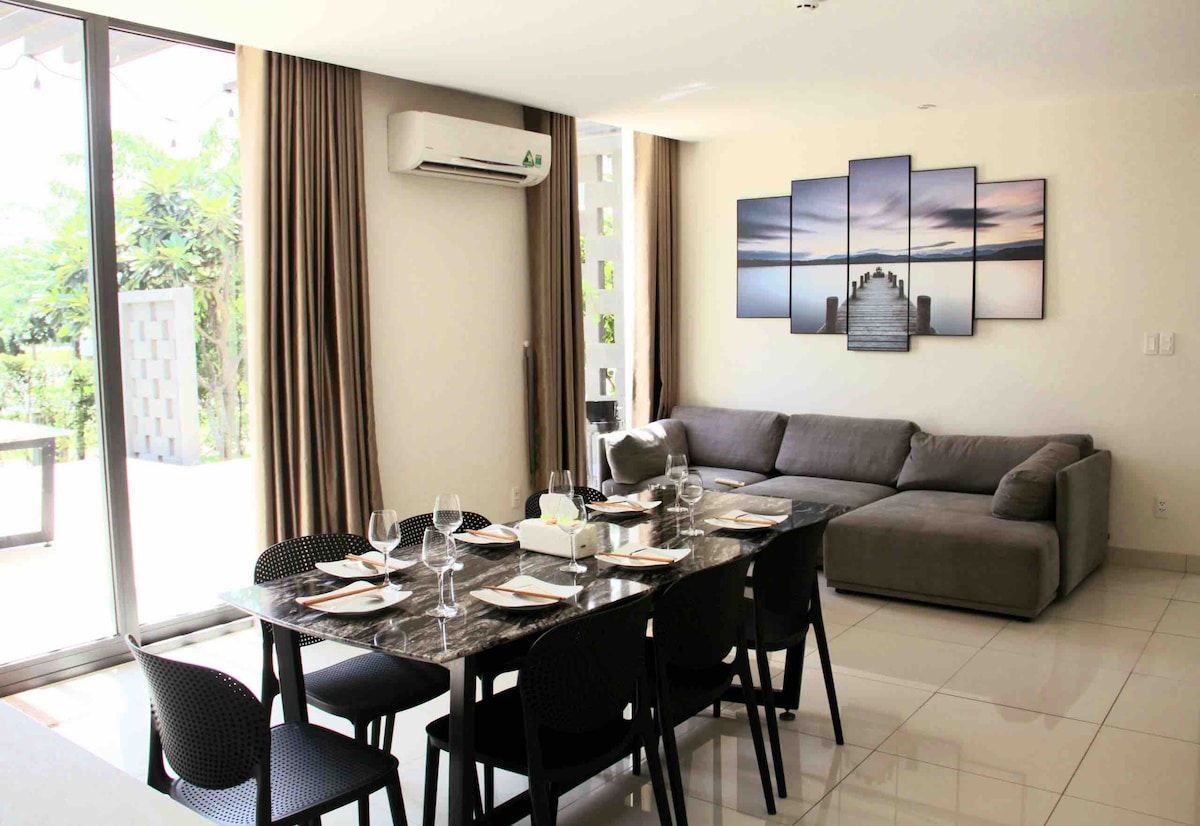An open dining area features a large table set for dining, complemented by a spacious grey sofa nearby. Large glass doors allow natural light to fill the room, while a decorative wall art piece above the sofa adds a touch of elegance.