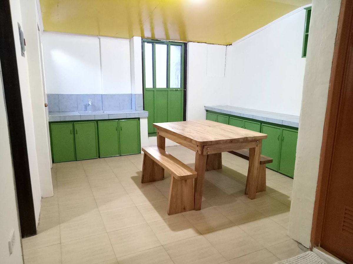 A light-filled dining area features a wooden table with matching benches, positioned on tiled flooring. Green cabinets line the walls, complementing the bright yellow ceiling. Large windows allow natural light, enhancing the space's openness and functionality.