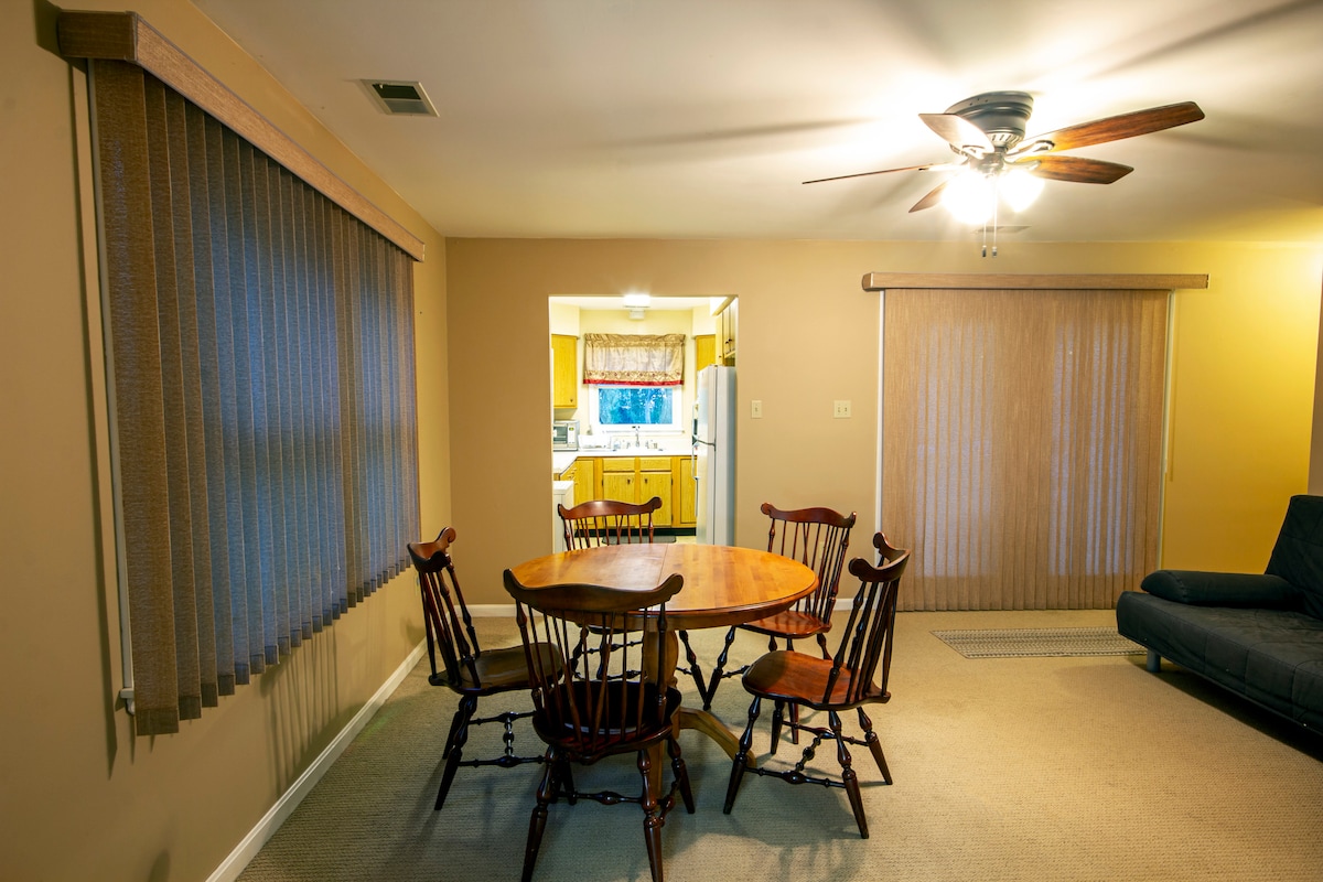 A dining area is highlighted by a wooden table surrounded by six wooden chairs. Natural light enters through large windows covered by vertical blinds. In the background, an adjacent kitchen is visible through an open doorway.