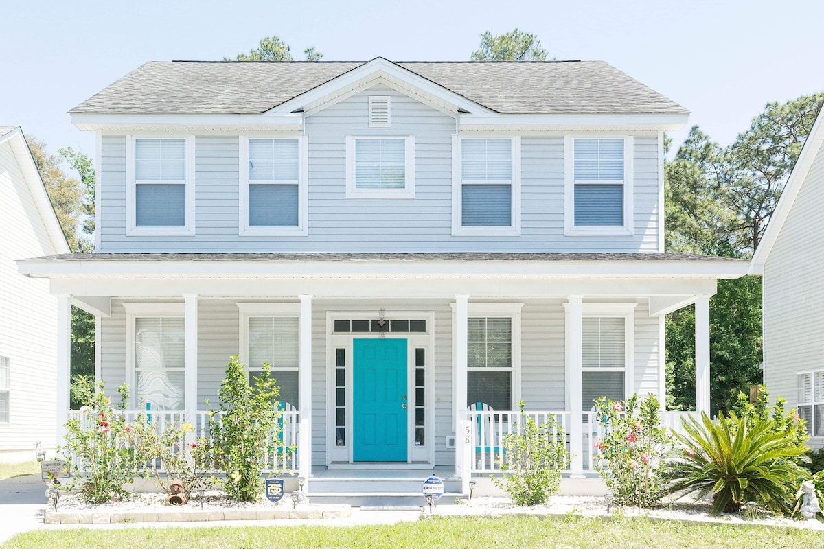 A two-story house is showcased, featuring light gray siding and a vibrant turquoise front door. The inviting porch is supported by white pillars and is adorned with planters. Lush greenery, including shrubs and palm plants, complements the front yard.