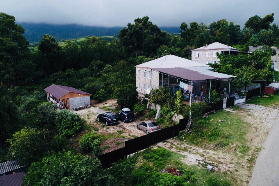 An aerial view captures a two-story house surrounded by abundant greenery. Several vehicles are parked in a designated area, while a smaller structure is visible nearby. Lush trees frame the scene, highlighting the natural setting of the suburban location.