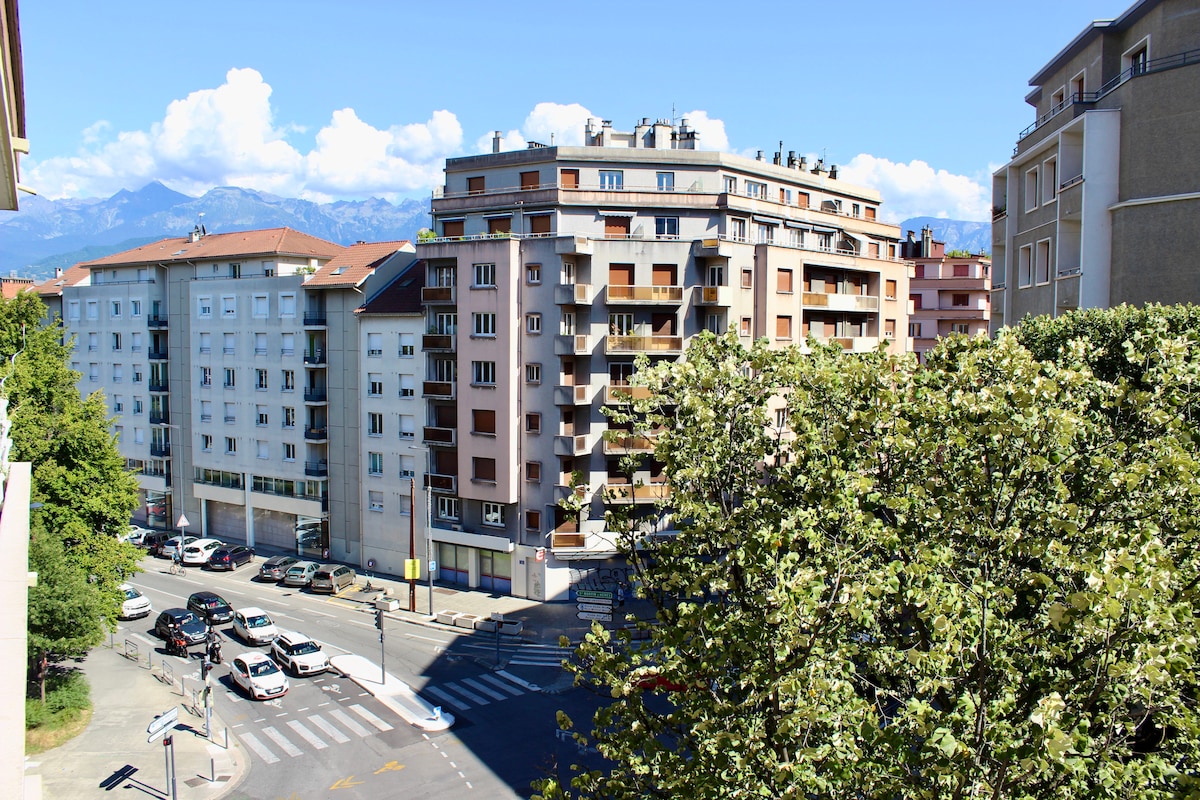 A view of the surrounding neighborhood displays multi-story buildings and tree-lined streets. The scene features an intersection with parked cars and pedestrians. The backdrop includes distant mountains under a clear blue sky, creating a tranquil urban landscape.