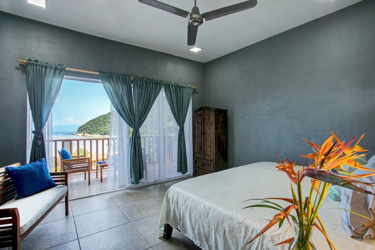 A spacious bedroom features a king bed with white linens and a wooden wardrobe against a dark wall. Two large windows are dressed with sheer curtains, allowing natural light and a view of the balcony, which offers a glimpse of the ocean.