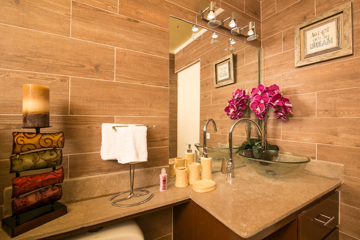 A modern bathroom features warm wood paneling and a glass vessel sink. Decorative candles and neatly arranged skincare items are positioned on the counter. An orchid plant adds a touch of greenery, enhancing the serene ambiance of the space.