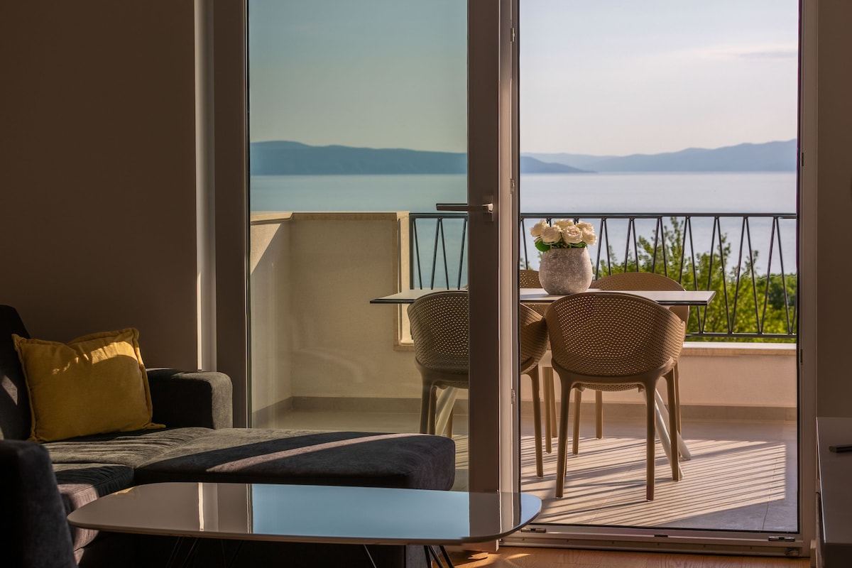A balcony with a dining table and chairs is visible through glass doors, offering views of the sea and distant hills. Natural light highlights a cozy seating area with a dark sofa and a coffee table set against a neutral wall.