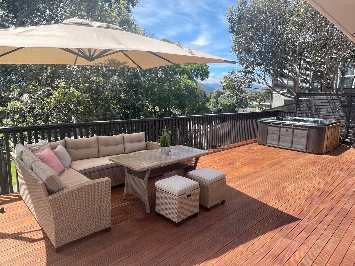 A spacious outdoor deck features comfortable seating arranged around a central table, shaded by large umbrellas. A spa pool is positioned nearby, surrounded by a wooden railing and mature trees, providing a private and relaxing atmosphere.
