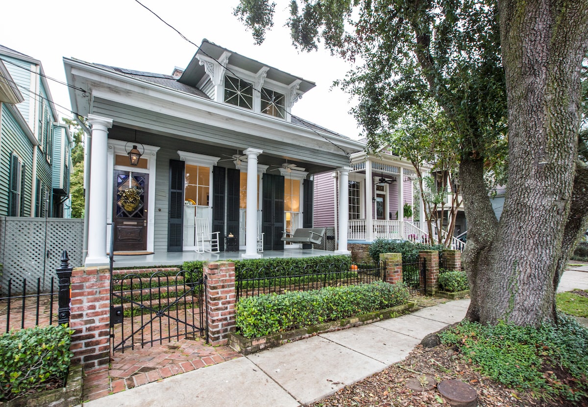 A charming two-story house features classic porch columns and a welcoming entrance. Lush greenery surrounds the brick walkway and wrought-iron fence. The façade showcases a blend of modern and traditional architecture, with adjacent homes reflecting the character of the neighborhood.