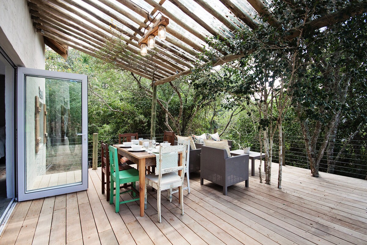 An outdoor seating area features a wooden deck surrounded by trees, offering a natural ambiance. A large dining table is complemented by various seating options, including woven chairs and colorful accents. Soft lighting is provided by overhead fixtures, enhancing the inviting space.