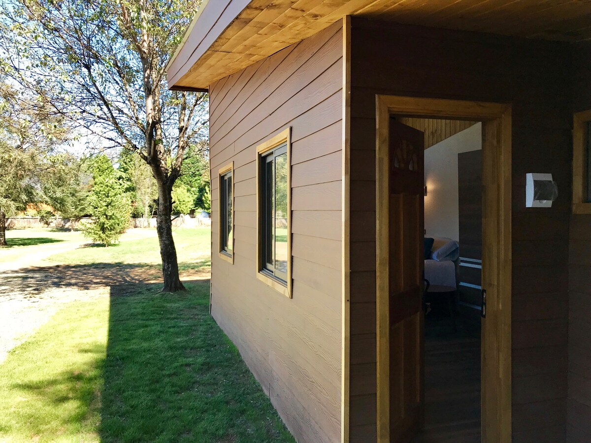 The exterior of a wooden cabin is shown, featuring a brown facade with three windows arranged symmetrically. Natural light filters in, enhancing the inviting entrance of the cabin. The surrounding area includes green grass and trees creating a serene mood.