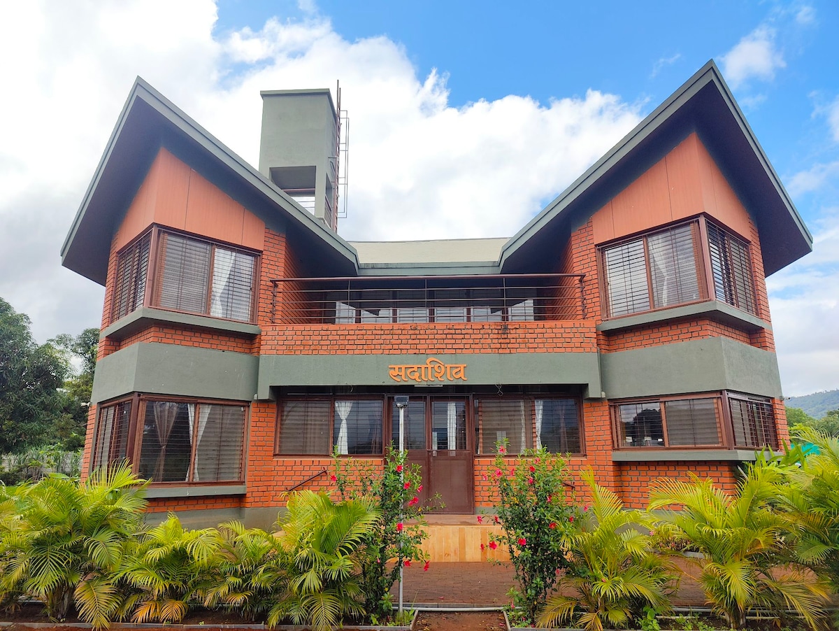 A modern two-story structure features a combination of brick and wood paneling. Large windows allow for natural light to fill the interior while providing views of the surrounding landscape. Lush greenery and flowering plants frame the entrance, enhancing the welcoming appearance of the home.