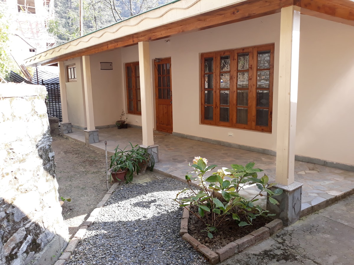 A charming cottage exterior is shown, featuring a covered porch with wooden trim and multiple large windows. A well-maintained garden area includes green plants and gravel pathways, while the stone wall and ground provide a natural setting. Sunlight filters through the surrounding trees.