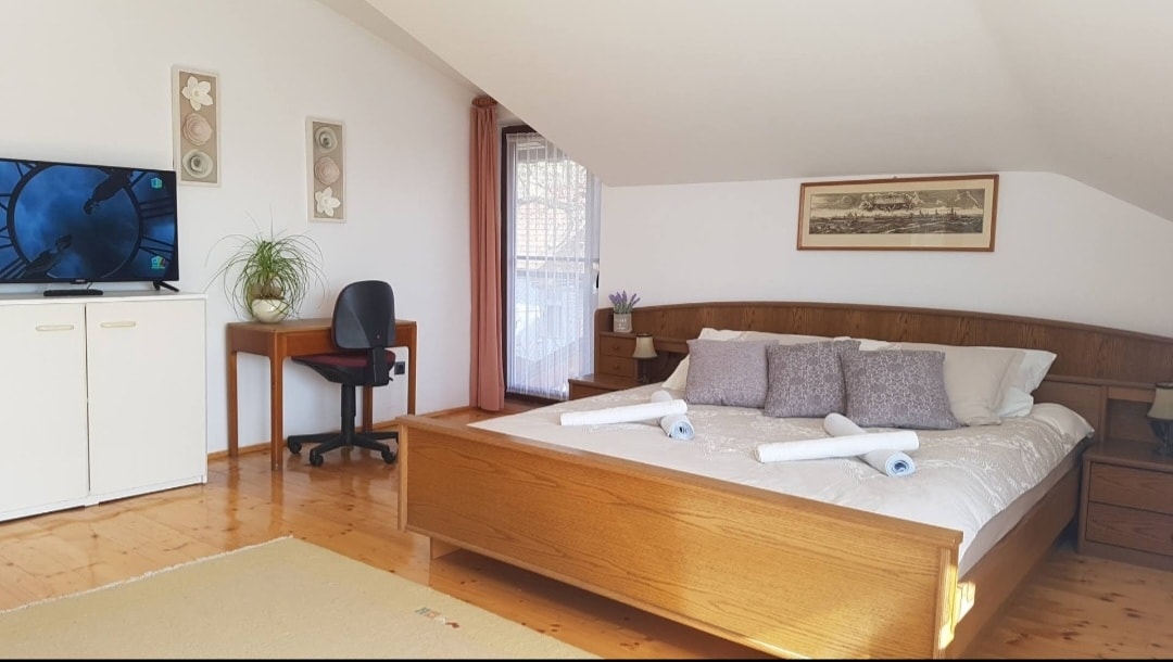 A spacious bedroom features a wooden bed adorned with decorative pillows and neatly arranged towels. A television is mounted on the wall, alongside a small desk and chair. Natural light filters through a window, complemented by light-colored curtains, enhancing the room's bright ambiance.