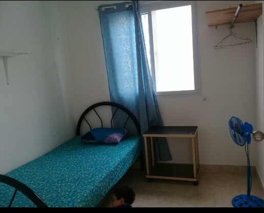 A simple sleeping area is depicted, featuring a single bed with a blue floral coverlet. A window allows natural light, while a blue curtain adds a pop of color. A small wooden side table and a fan are also visible, providing functional furnishings.