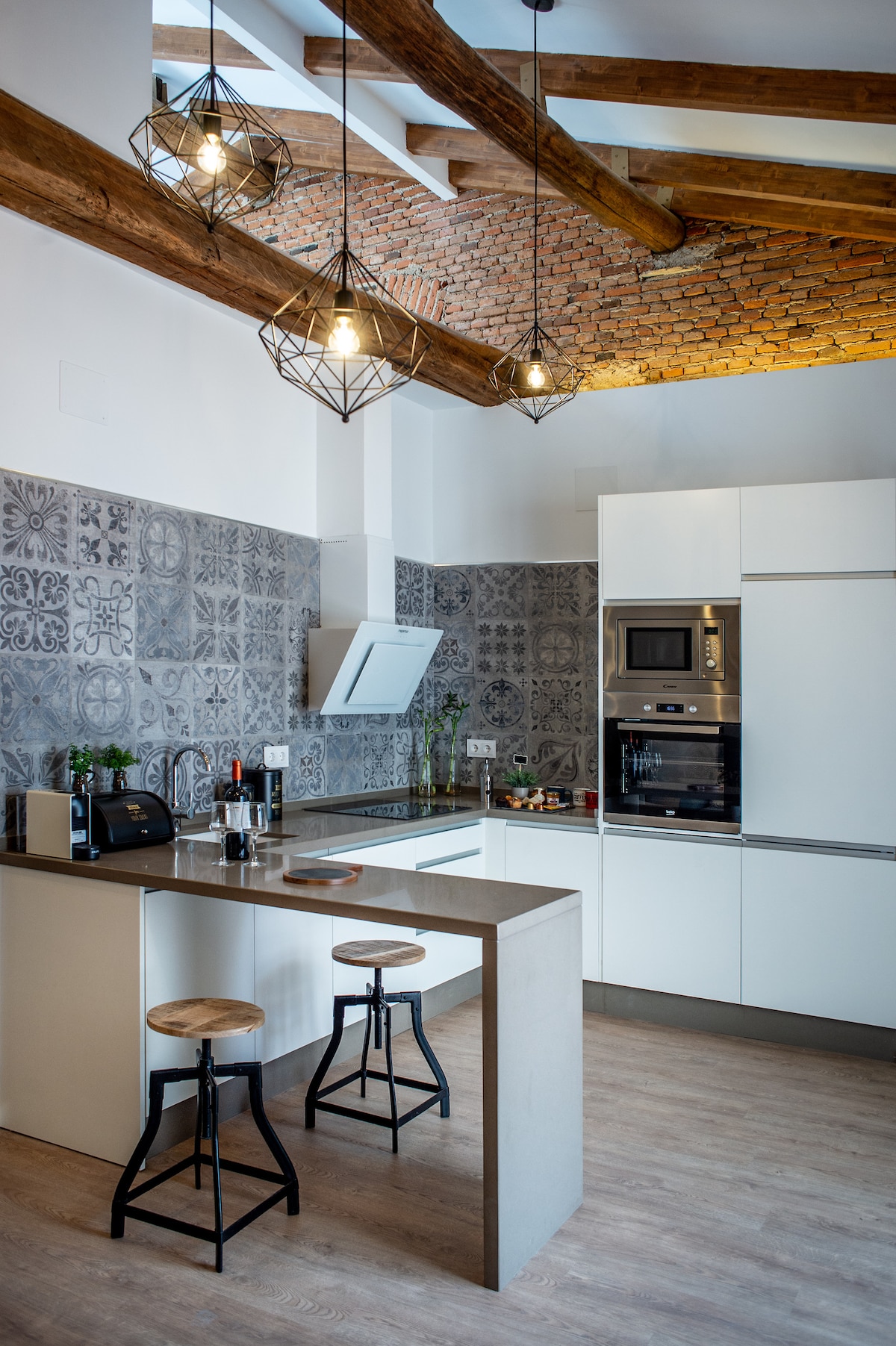 The kitchen showcases a blend of modern design and rustic charm, featuring open wood beams and patterned tile backsplash. Two sleek stools are positioned at a minimalist bar, while contemporary appliances and decorative plants complete the functional layout.