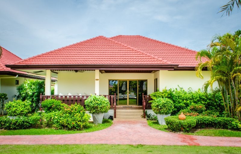 The exterior of a bungalow is presented, featuring a red tiled roof and a welcoming entrance. Lush green plants and hedges surround the front, while a path leads up to the entrance, enhancing the inviting look of the property.