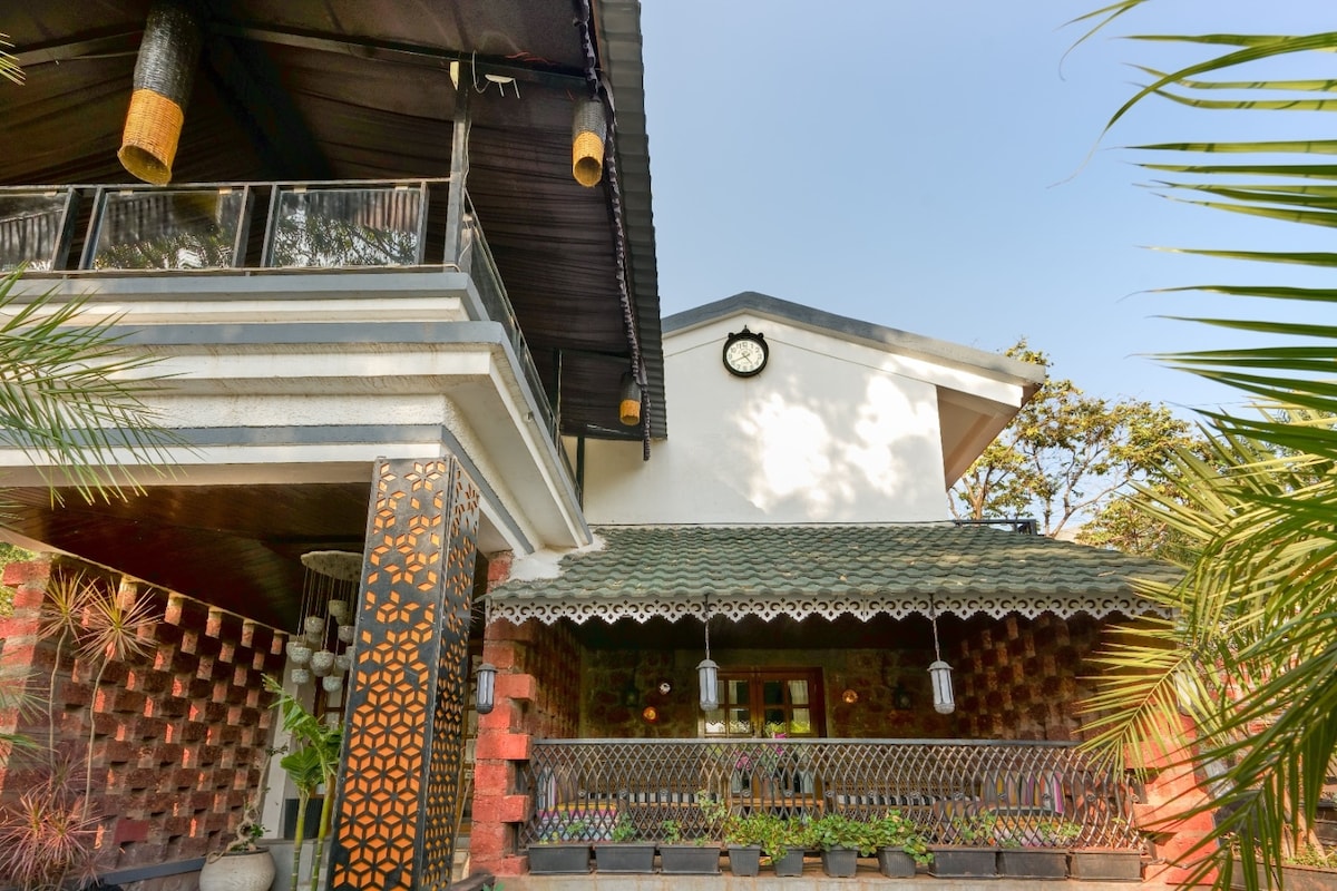 The exterior of Azalea Villa is displayed, featuring a blend of modern and traditional architecture. A large clock adorns the upper wall, while decorative lanterns hang from the ceiling. Lush greenery surrounds the verandah, accentuated by potted plants along the railing.