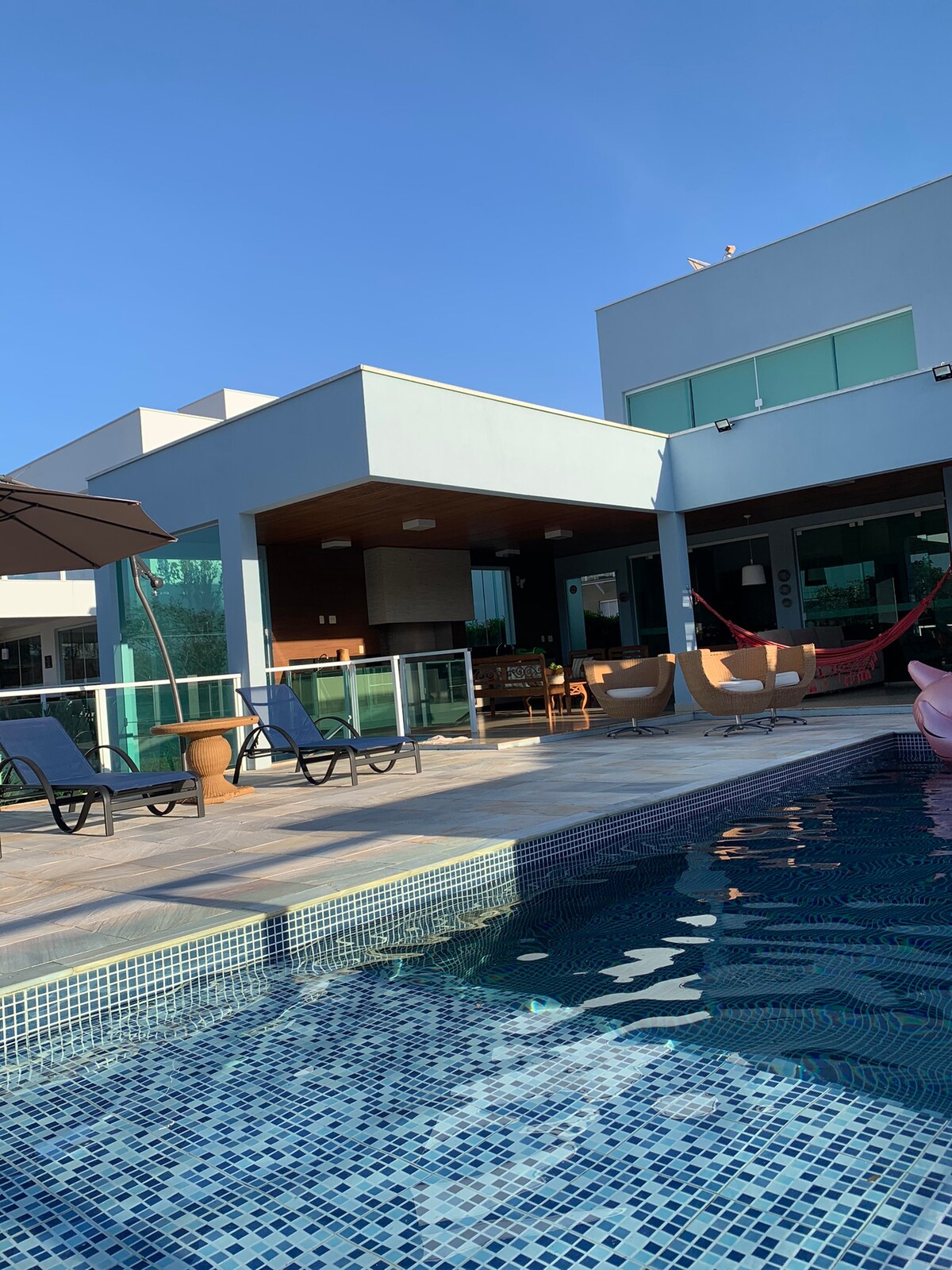 A modern swimming pool is surrounded by lounge chairs and an umbrella, leading to a spacious deck. The house features large windows and a seamless design that connects indoor and outdoor spaces. A dining area is visible under a shaded section, creating an inviting space.