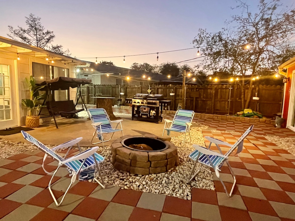 An outdoor patio area features a circular stone fire pit surrounded by four striped lounge chairs. Soft string lights hang above, creating a warm ambiance. A grill is positioned nearby, and a swing chair is visible in the background, contributing to a relaxed atmosphere.