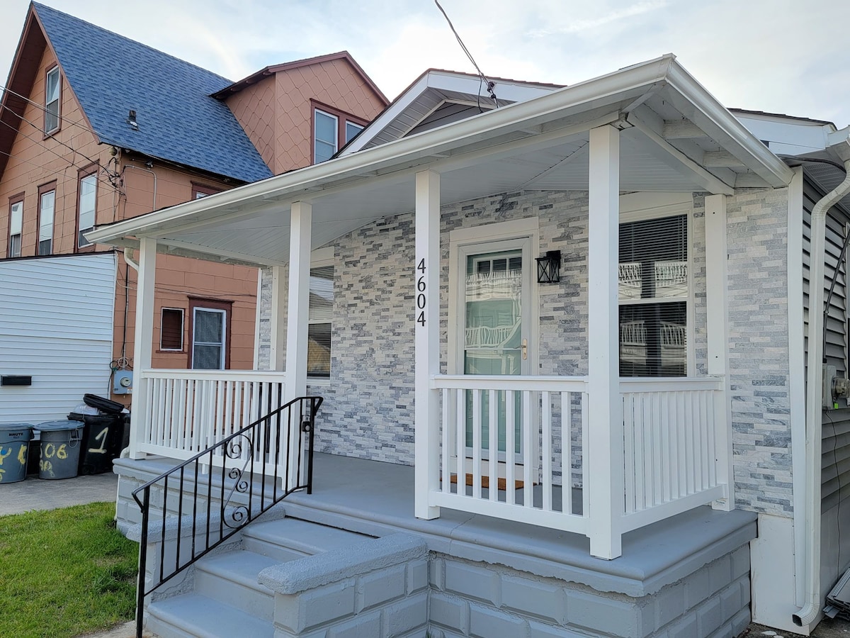 A front porch is shown, featuring white railings and steps leading to a glass front door framed by a light fixture. The exterior wall displays a combination of light stone and grey siding, contributing to the entrance's distinctive appearance.