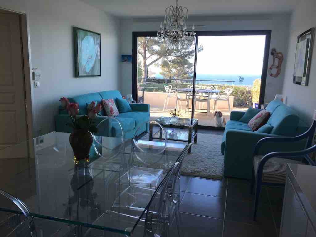 A light-filled living space features turquoise furnishings, including a sofa and chairs. A glass table reflects natural light, complementing the sea view visible through expansive sliding doors. A dining area transitions to an outdoor terrace, creating a seamless blend between indoor and outdoor living.