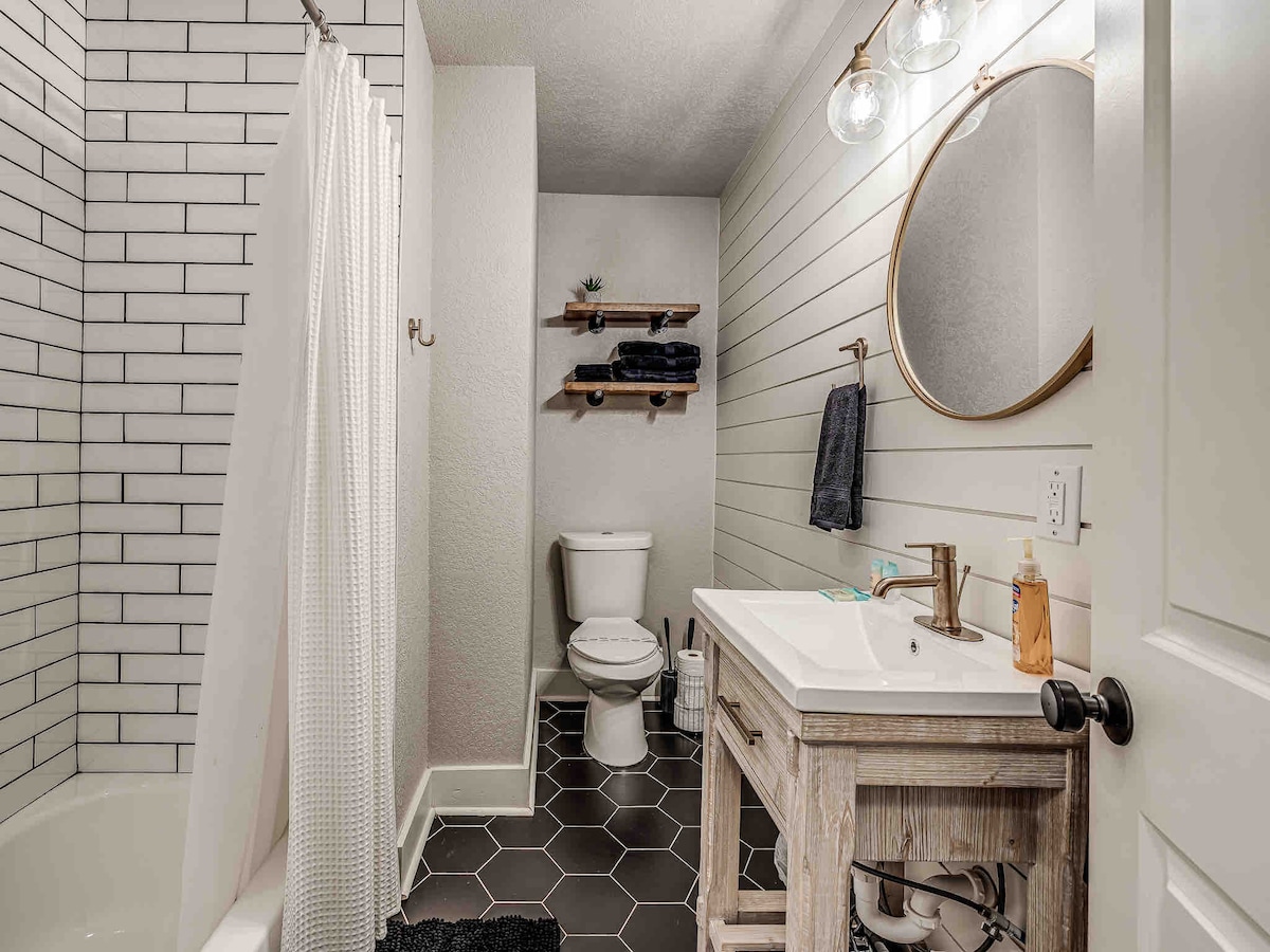 A modern bathroom features a tiled shower area with a white curtain. A wooden vanity holds a round mirror and essential toiletries. Dark hexagonal tiles contrast with light walls, while shelves hold neatly arranged towels.