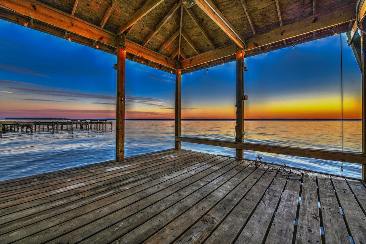 A wooden deck with a rustic ceiling offers an unobstructed view of the water at sunset. The soft colors of the horizon blend into the tranquil surface of the lake, while string lights gently illuminate the space, enhancing the serene atmosphere.