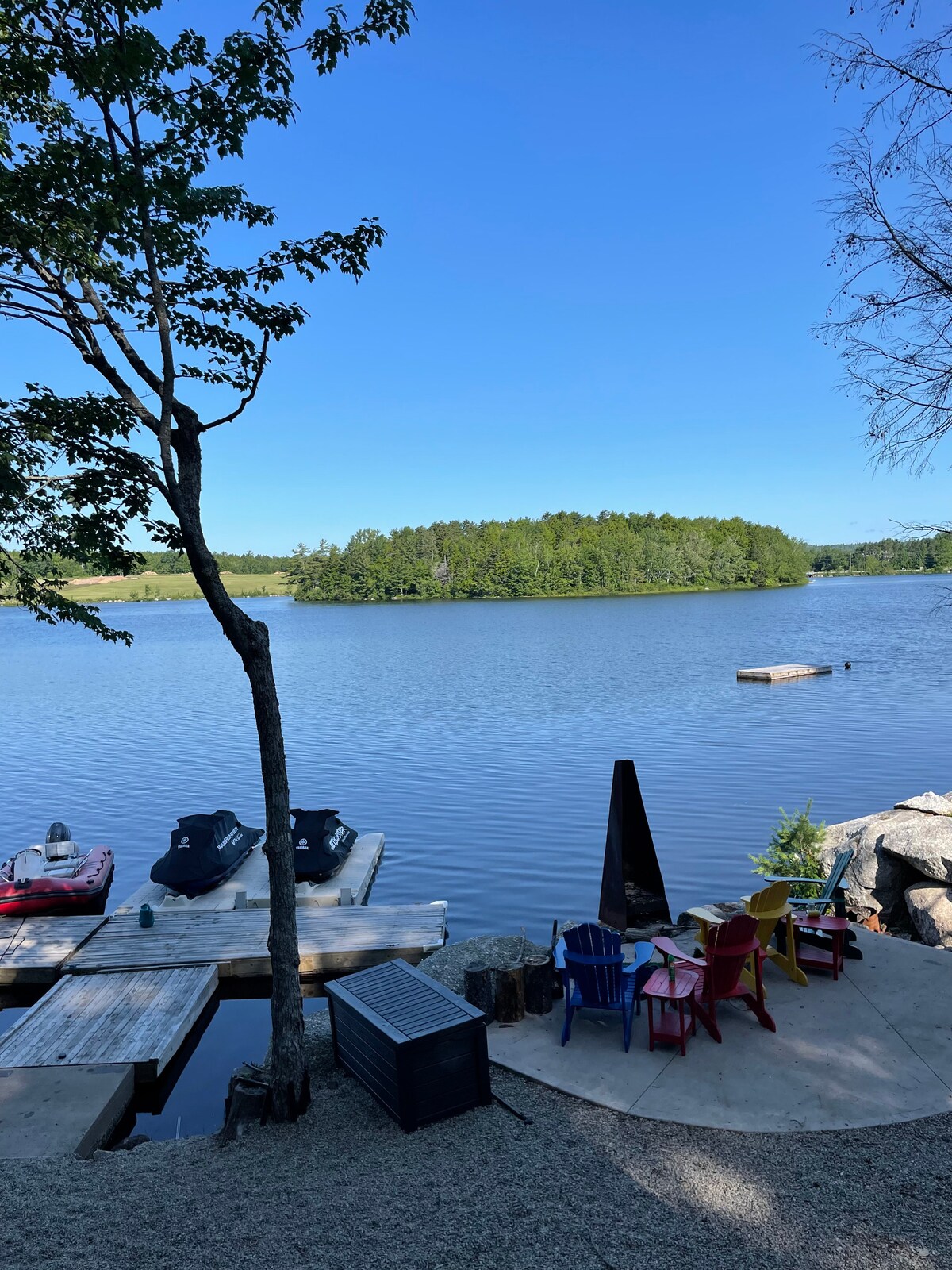 A serene lake view is displayed, featuring calm waters and lush greenery across the water's expanse. Colorful Adirondack chairs are arranged around a tiered stone fire pit area, while kayaks are secured at the dock. A single swim dock is visible on the water.
