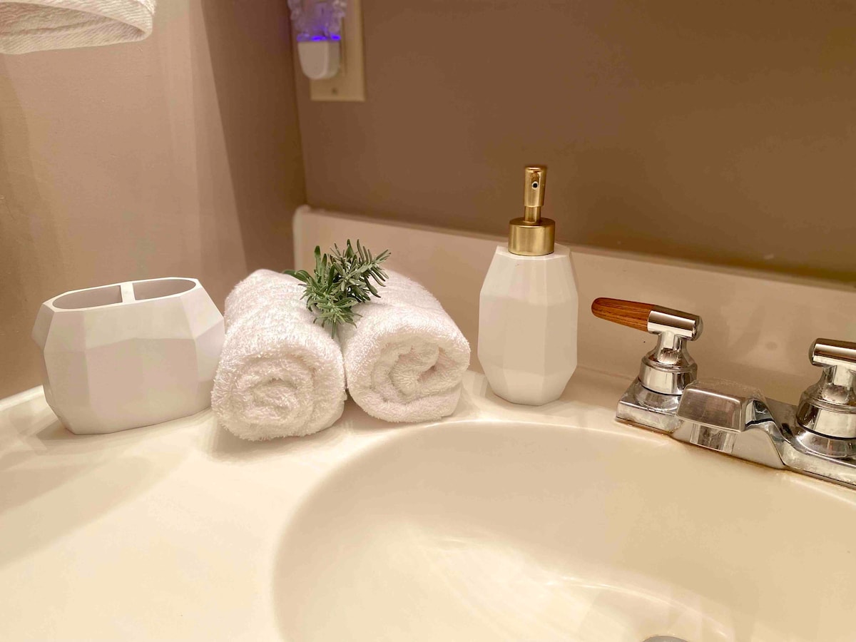 A clean countertop features two neatly rolled white towels accompanied by a small green accent. A white ceramic container and a white soap dispenser with a wooden handle are also present, along with polished chrome faucets.