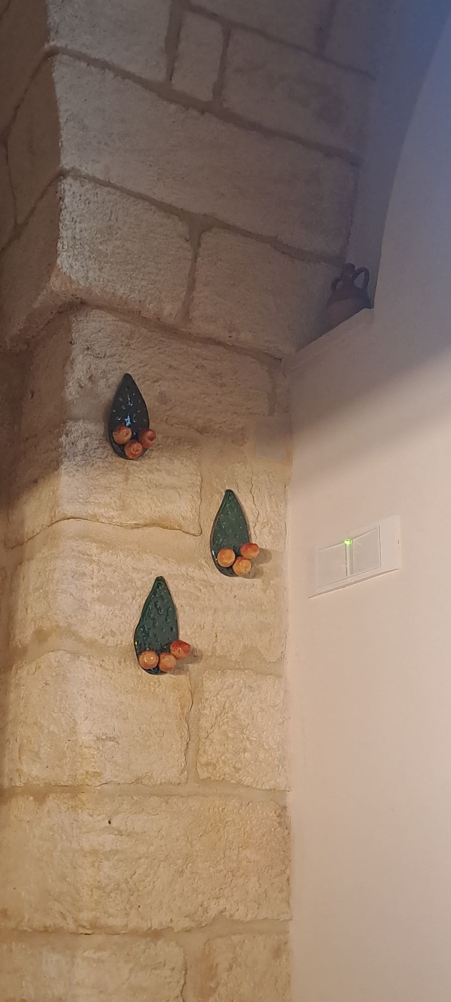 A corner of the wall features three decorative elements resembling leaves adorned with small orange accents. The textured stone of the wall contrasts with the colorful decor, emphasizing the natural materials used in the home. A light switch is visible nearby.