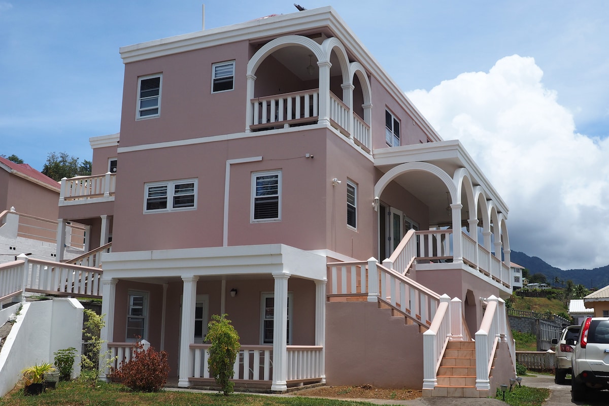 A two-story building with a light pink exterior is featured, characterized by multiple arched balconies and large windows. A staircase leads to a welcoming entrance, complemented by surrounding greenery and a clear blue sky.