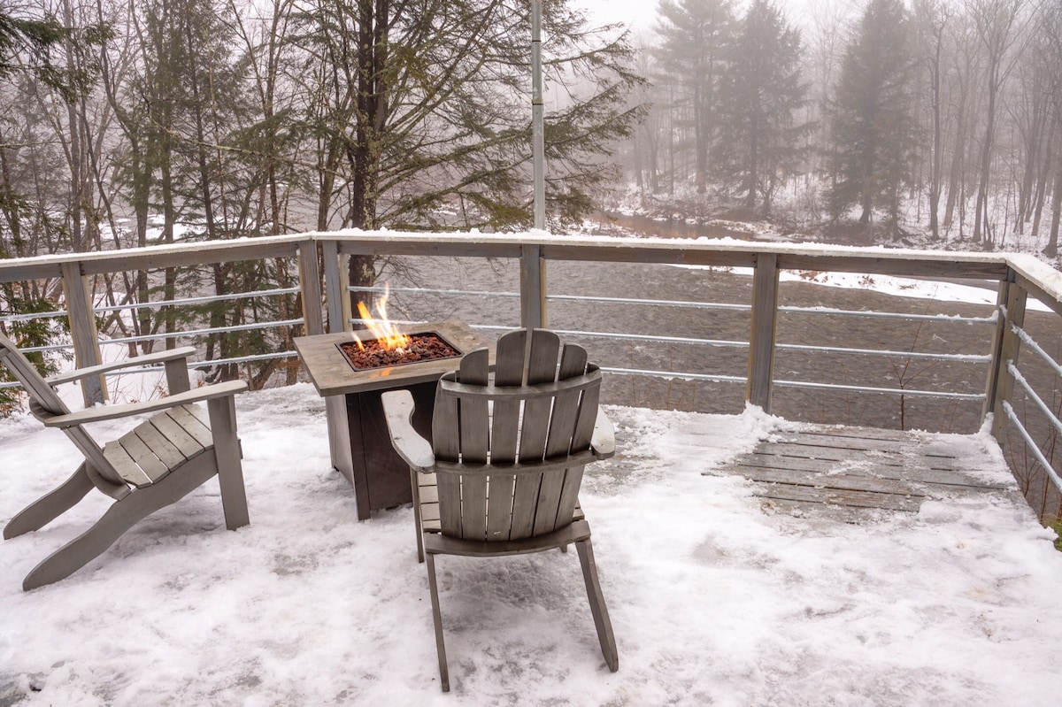 An outdoor deck is depicted, featuring two adirondack chairs positioned beside a small table with a gas fire pit. Light snowfall covers the deck, creating a serene winter scene overlooking a foggy river and surrounding trees.