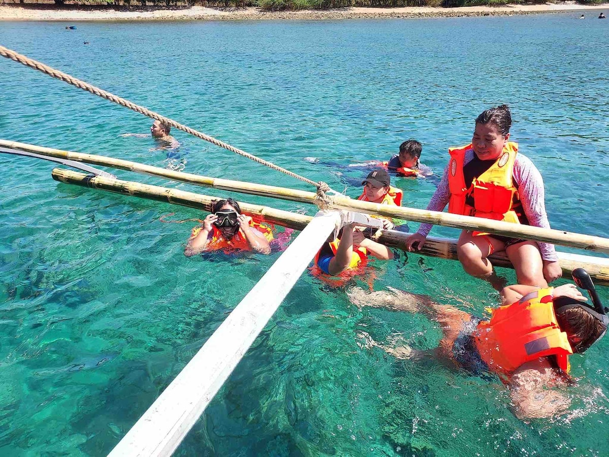 Individuals are seen engaging in a water activity while wearing bright orange life jackets. Some are sitting on a wooden structure, while others are partially submerged in clear, turquoise water. Sunlight sparkles on the surface, enhancing the lively setting.