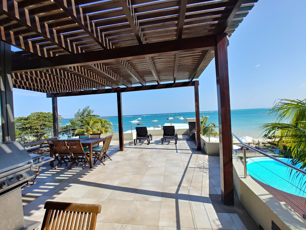A spacious outdoor terrace is highlighted by a wooden pergola, overlooking the turquoise ocean. A dining area is set with a large table and wooden chairs, while two lounge chairs are positioned near the edge, providing stunning views of the beach and water.