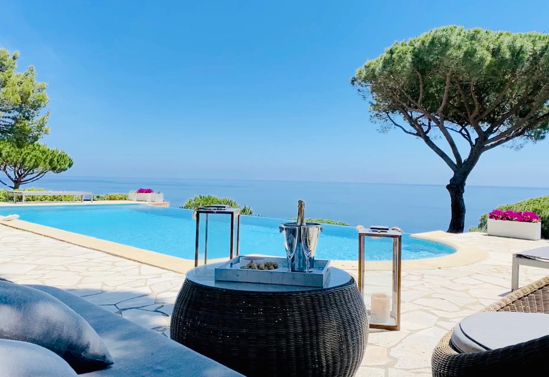 An infinity pool extends toward the horizon, reflecting clear blue skies and calm waters. Comfortable seating is arranged around a round table, featuring a cooler and decorative items. Lush trees and vibrant flowers enhance the serene view, providing a tranquil outdoor setting.
