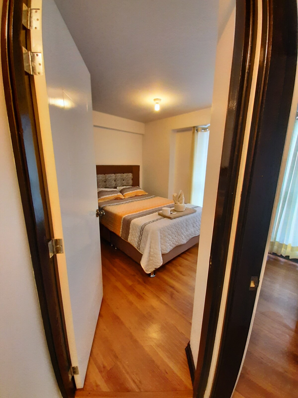A bright bedroom is visible through an open door, featuring a spacious bed with neatly arranged bedding. Soft lighting illuminates the room, and large windows with sheer curtains allow natural light to fill the space, creating a welcoming atmosphere.