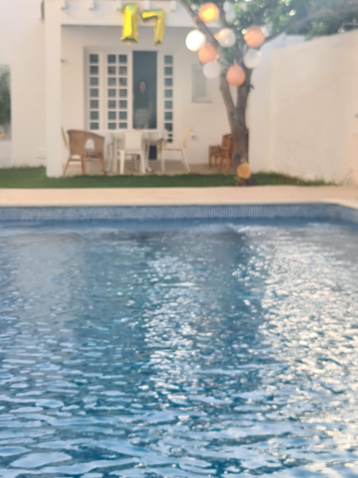 A clear swimming pool reflects the surrounding greenery, with a well-maintained garden behind it. In the background, a dining area with chairs and a table is visible, framed by soft lights hanging from nearby trees.