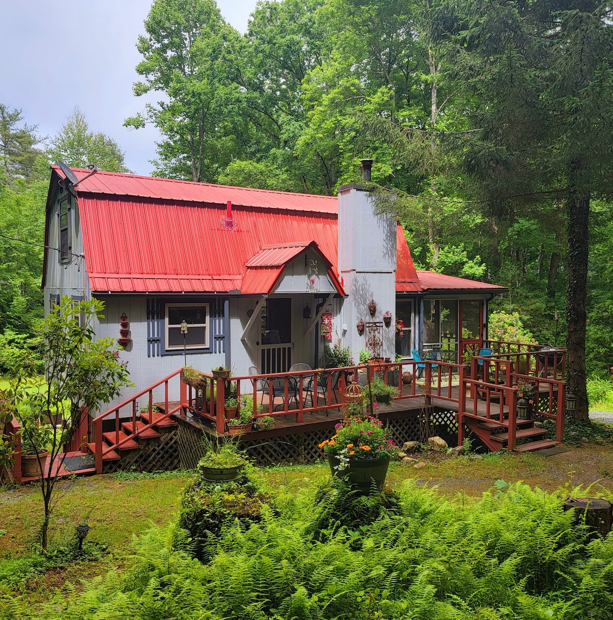 A charming cottage is surrounded by lush greenery, featuring a striking red metal roof. The entrance is framed by a wooden deck with flower pots and decorative elements. Tall trees provide shade, creating a serene atmosphere.