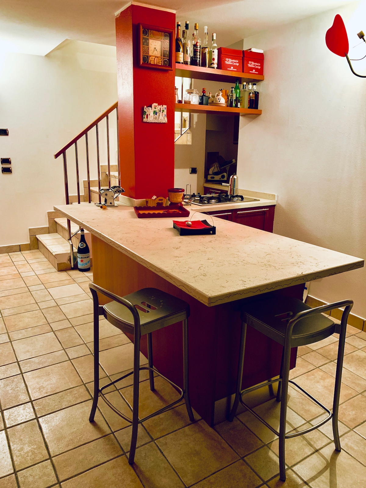An open kitchen area features a stone countertop with two metal stools. Above, shelves display various bottles. The room is lit by a wall-mounted light fixture, with a staircase visible in the background, leading to other areas of the home.