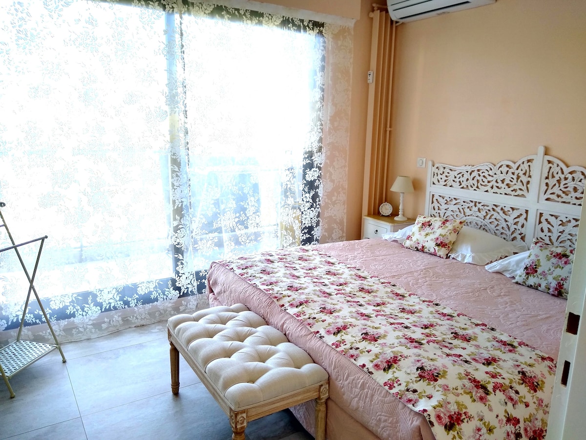 A cozy bedroom features a spacious bed adorned with floral bedding, complemented by a decorative bench at the foot. Large glass doors with lace curtains allow soft light to fill the space, enhancing the tranquil ambiance. A bedside lamp is visible on a small table.