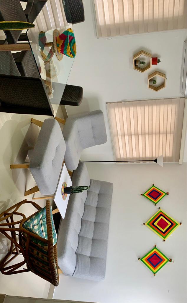 A modern living area features a grey sofa and ottoman, complemented by a rattan chair. A glass dining table is set nearby. Colorful wall decorations add visual interest, while natural light filters through the window coverings, creating a bright and inviting space.