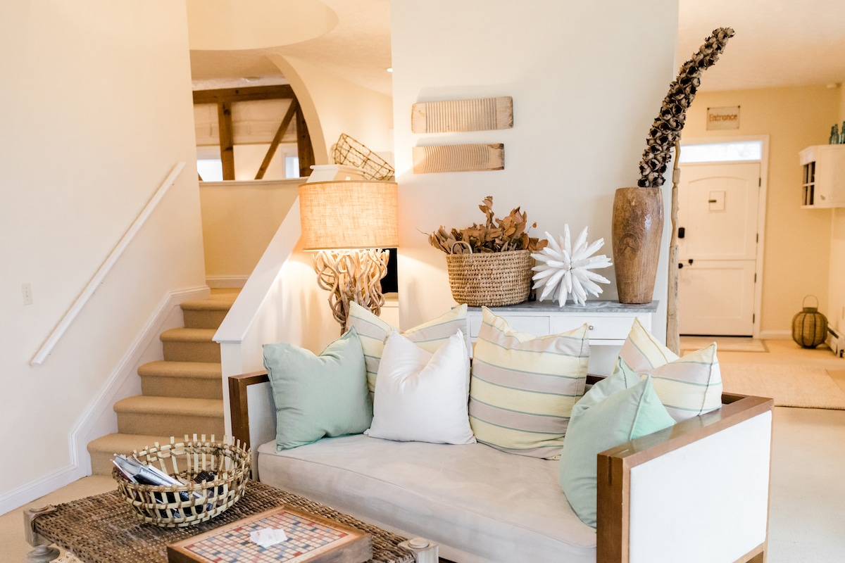 A bright and airy living area features a comfortable seating arrangement, showcasing plush pillows in various pastel colors. Natural light filters in, highlighting a woven basket filled with decorative elements. A staircase leads to the upper levels, enhancing the open layout of the space.