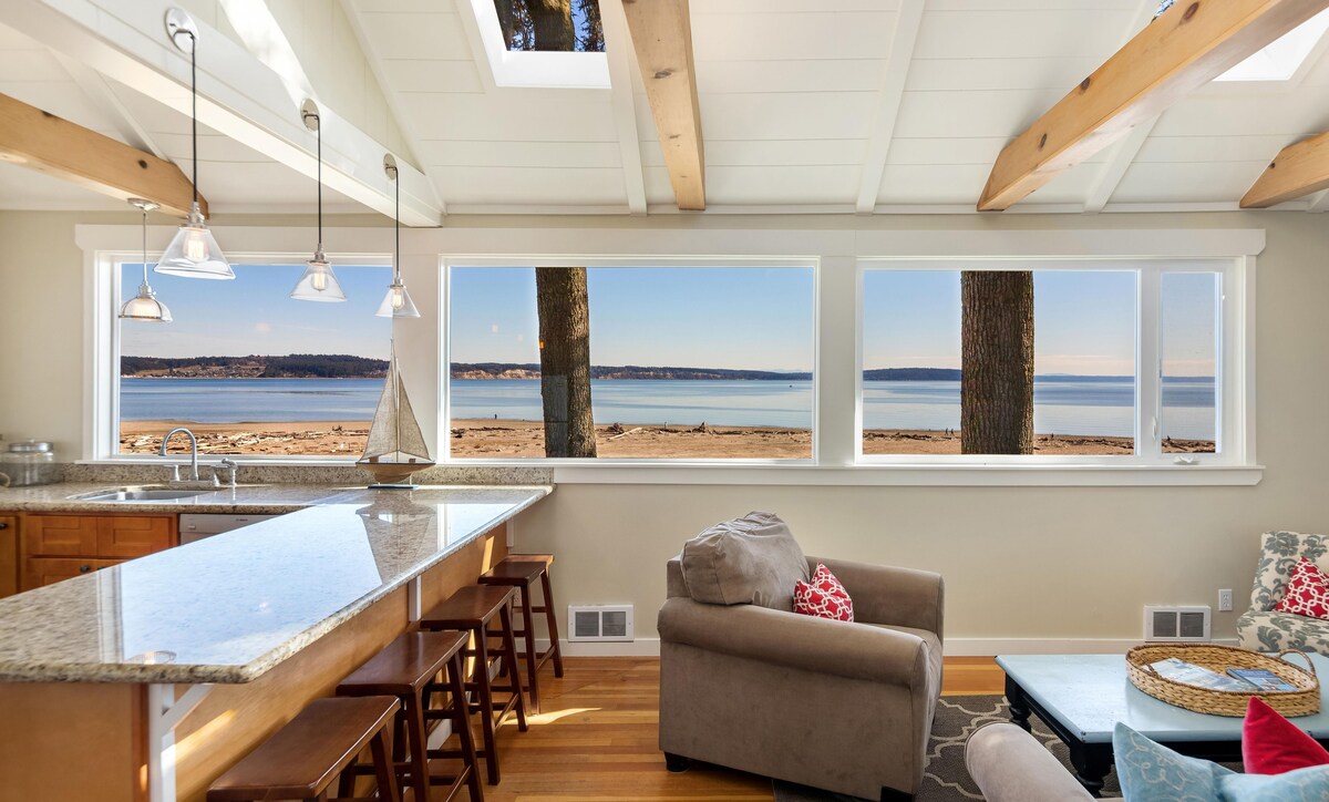The spacious kitchen features a granite countertop with bar seating for three. Large windows allow natural light to fill the room, offering views of the sandy beach and water beyond. Exposed wooden beams add a touch of warmth to the bright and airy design.