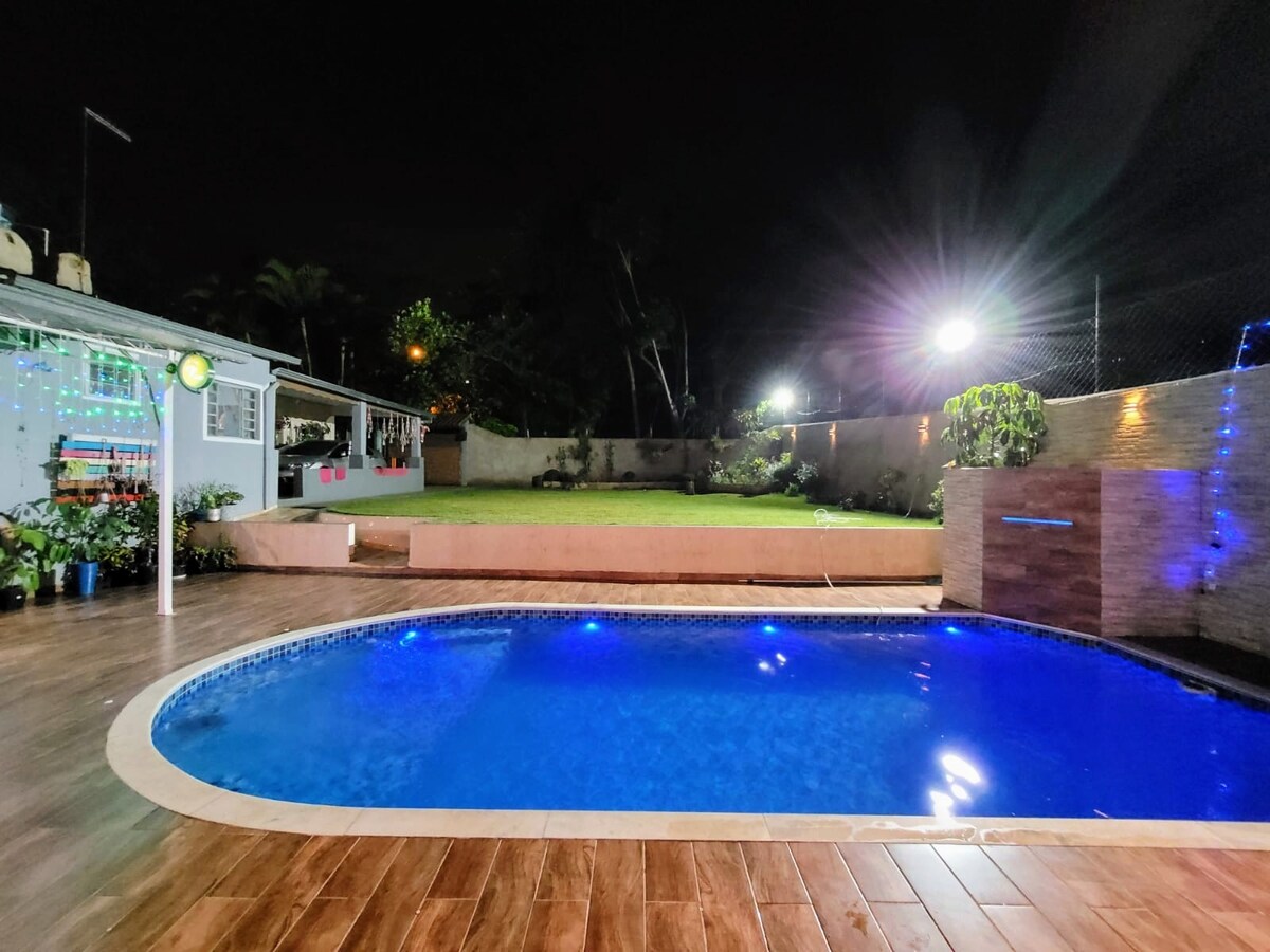 A tranquil outdoor space features a shimmering pool illuminated by soft blue lighting, surrounded by a wooden deck. Lush greenery is visible in the background, creating a calming atmosphere. Outdoor seating and decorative lights add to the inviting environment.