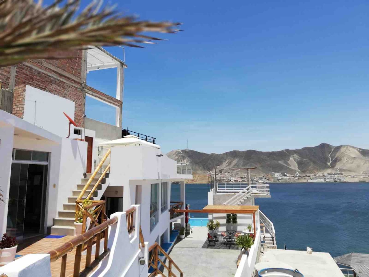 The image presents a view of a multi-level house on a waterfront, with stairs leading to upper terraces. Various outdoor areas offer seating, and a shared pool is visible. Natural light brightens the white exterior, complementing the surrounding landscape and ocean views.