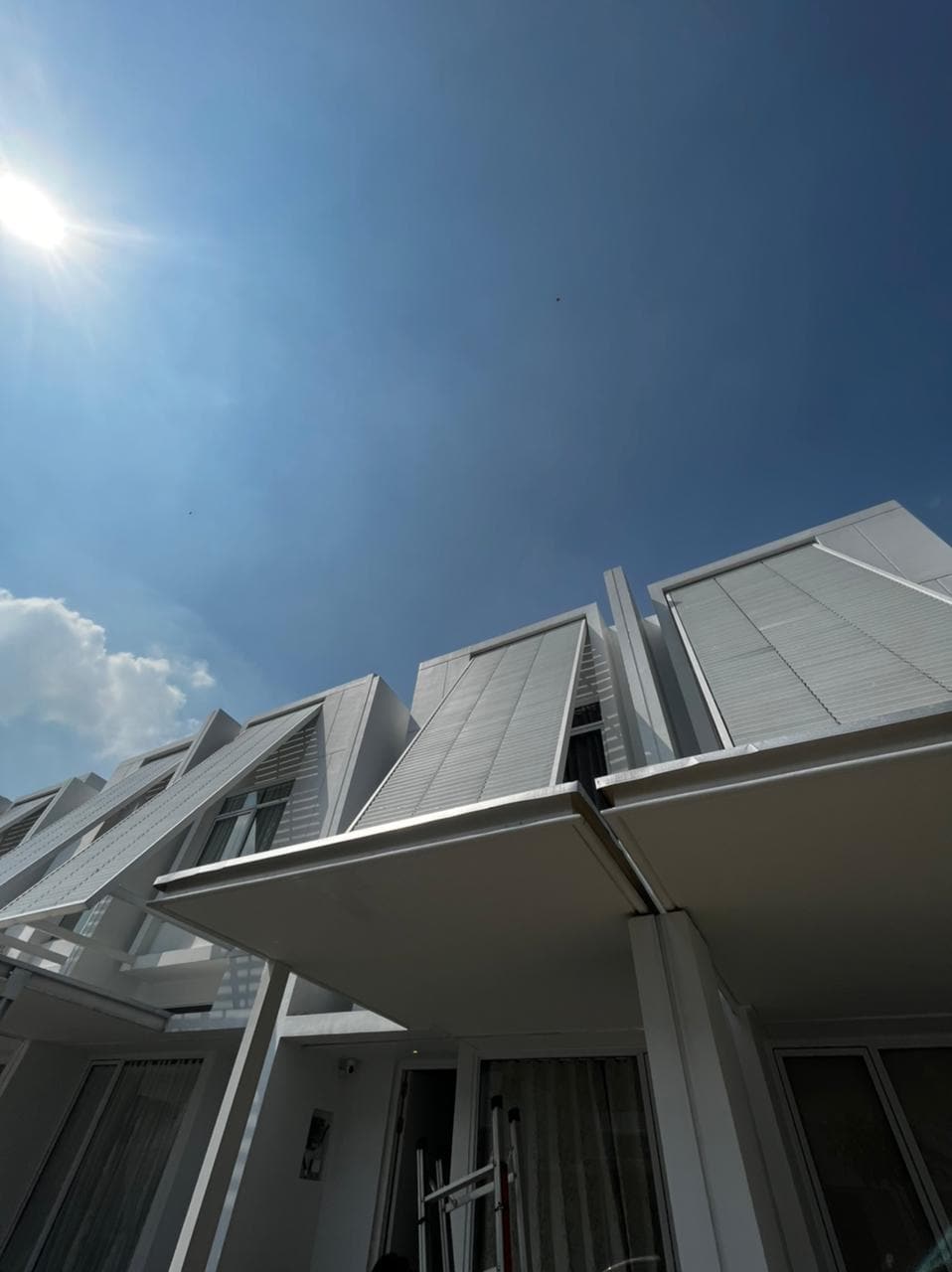 A modern architectural structure displays a sleek design with asymmetrical lines and large windows. The façade, painted in a light color, reflects the bright sky. Sunlight casts shadows on the exterior, enhancing the clean and contemporary look of the building.