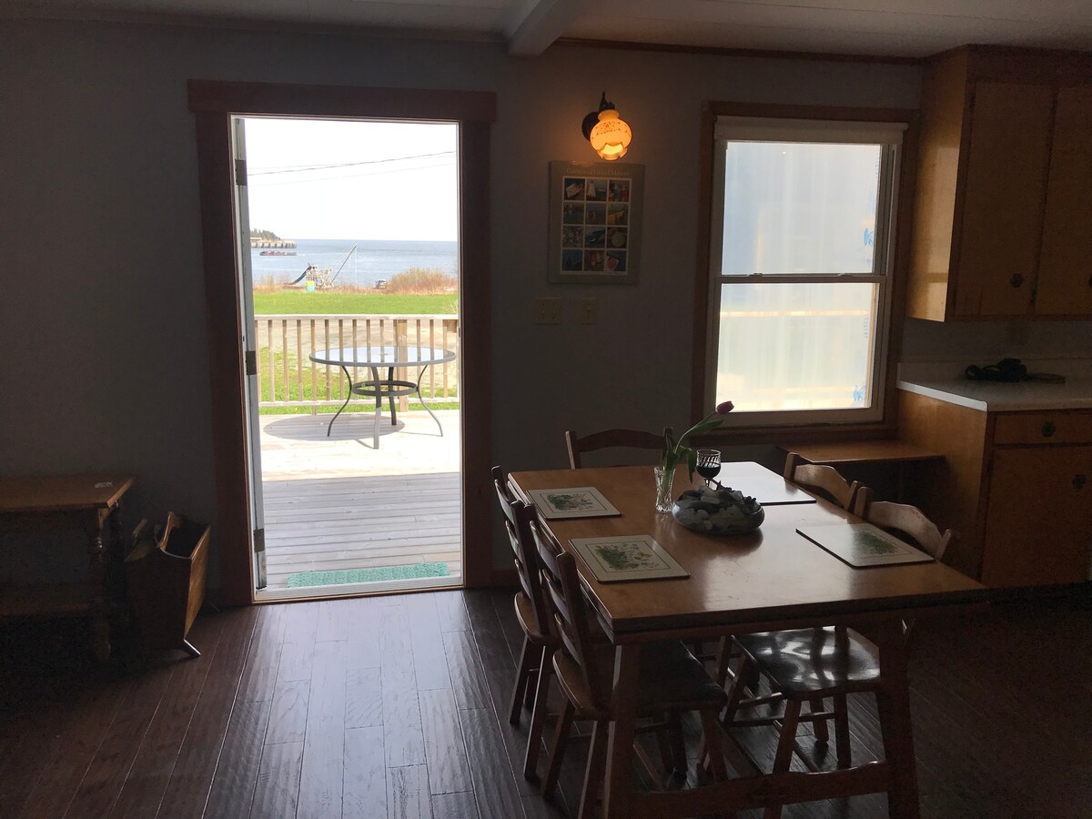 A cozy dining area is presented with a wooden table surrounded by chairs. A door opens to a deck with views of the ocean. Natural light floods the room, highlighting the simple cabinetry and inviting atmosphere.