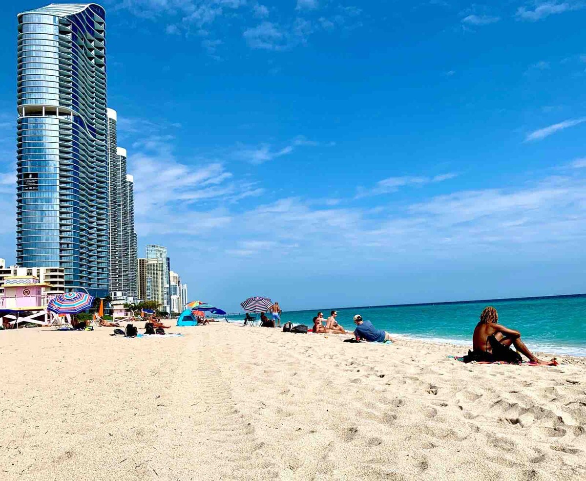The sandy beach is lined with colorful umbrellas and beachgoers enjoying the sunny day. Towering structures rise in the background, providing a contrast to the tranquil sea and clear blue sky. The shore offers a peaceful and spacious environment for relaxation.