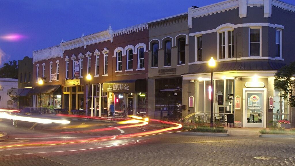 A row of charming commercial buildings is illuminated during twilight. Warm lights from storefronts create a welcoming atmosphere. The façade features a variety of architectural styles, with arched windows and contrasting colors. Light trails from passing cars add dynamic movement to the scene.