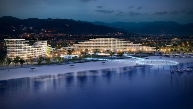An aerial view captures a luxury resort illuminated at night, showcasing modern architecture along the shoreline. The buildings are arranged in elegant formations, reflecting softly on the water, while the surrounding landscape features trees and pathways. The distant hills provide a serene backdrop.