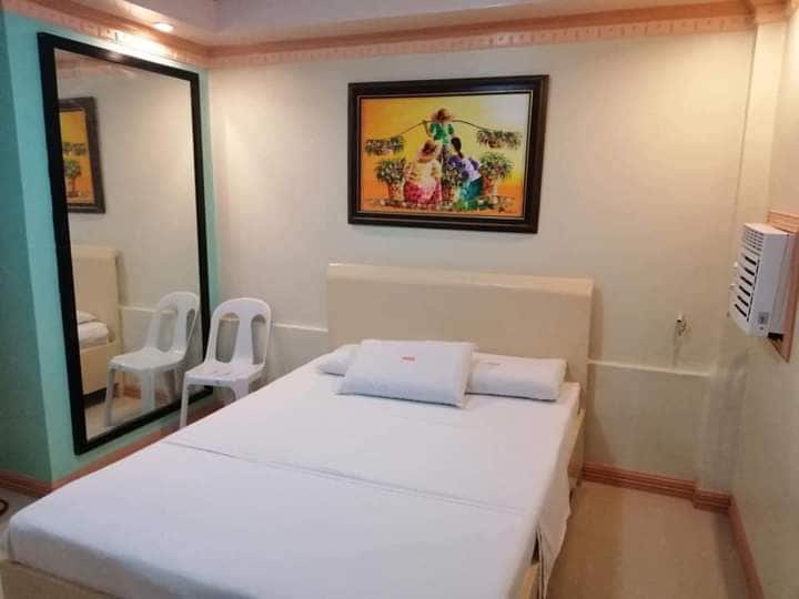A bright sleeping area is furnished with a spacious bed, complemented by a large framed artwork on the wall. A mirror reflects the room's layout, which includes two simple white chairs positioned beside the bed. Air conditioning is available for comfort.